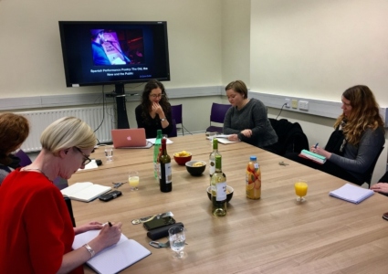 Seminar speaker delivering talk at a boardroom table with large display screen behind. Guest taking notes at same table with refreshments in the centre,