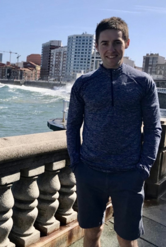 Ciaran Harty smiling wearing standing in front of a balustrade with the sea and high-rise building work behind