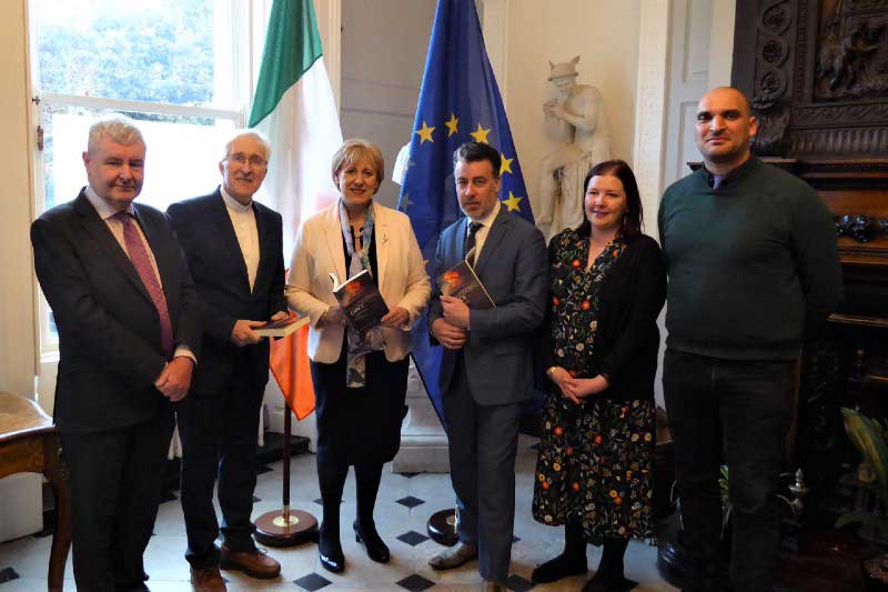 four men and two women in a row in front of a large window, with Irish flag, the European Union flag and a statue behind them