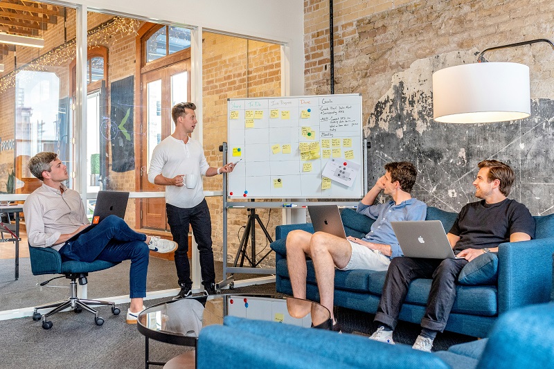 Four young professional men meeting and discussing in a bright informal space - three are seated and the other is posting post-it notes on a whiteboard. Photo by Austin Distel for Unsplash.